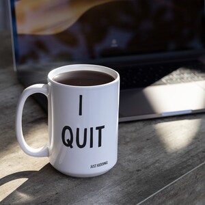 May include: White ceramic coffee mug with black text that reads "I QUIT JUST KIDDING". The mug is filled with coffee and is sitting on a wooden table next to a laptop.