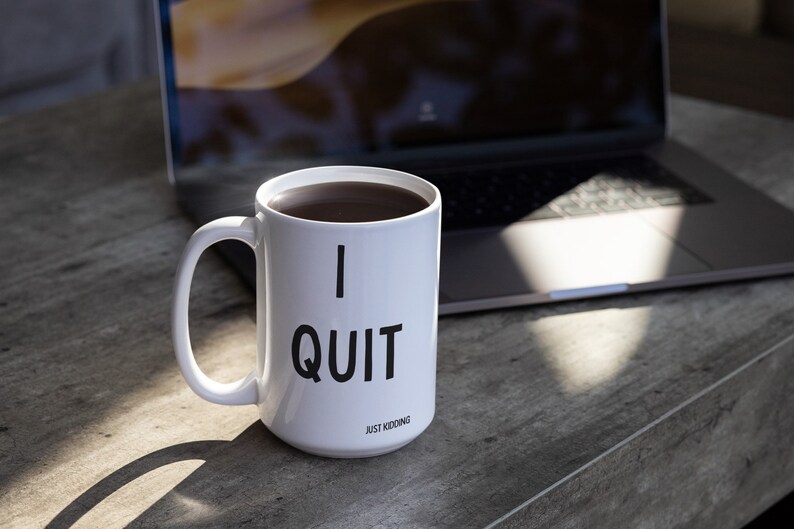May include: White ceramic coffee mug with black text that reads "I QUIT JUST KIDDING". The mug is filled with coffee and is sitting on a wooden table next to a laptop.