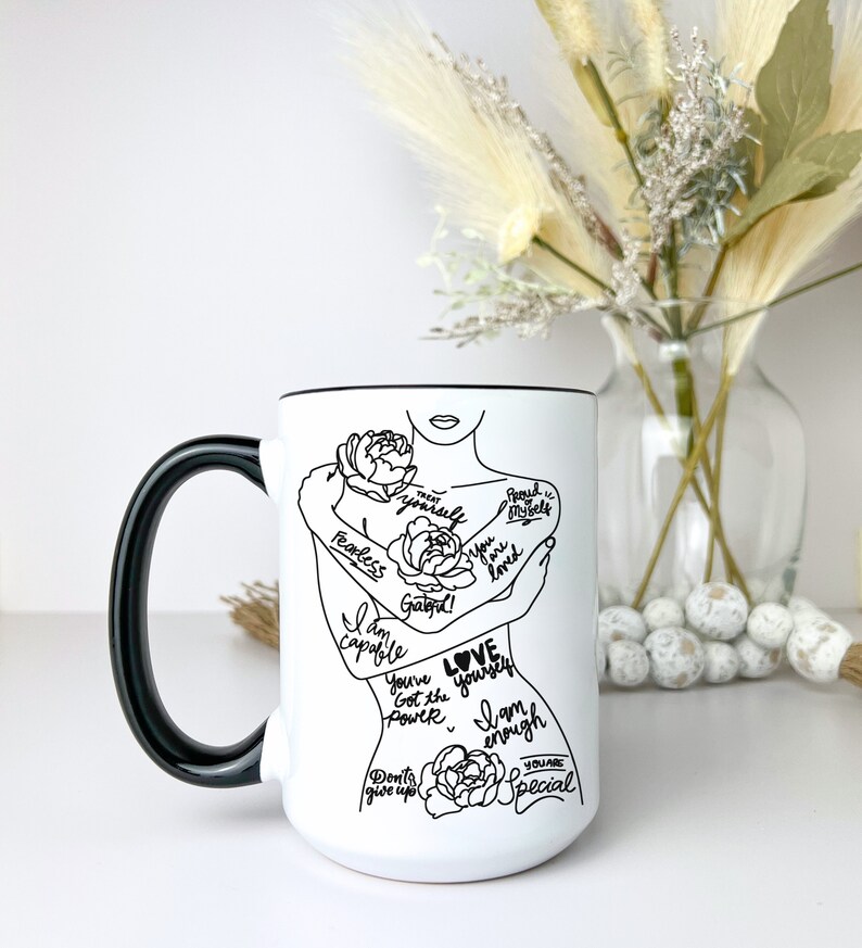 Puede incluir: Taza de cer&aacute;mica blanca con asa negra. La taza presenta un dibujo en l&iacute;nea negra de una mujer abraz&aacute;ndose a s&iacute; misma con las palabras "Love yourself" y otras afirmaciones positivas escritas en su cuerpo.