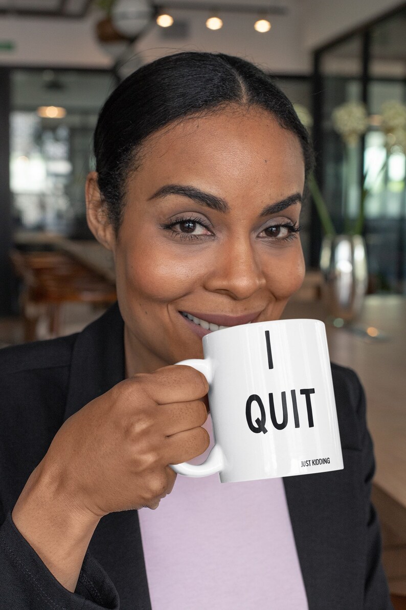 May include: A white ceramic mug with a black inscription that reads "I QUIT JUST KIDDING". A person is holding the mug and smiling.