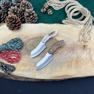 May include: Two small, handcrafted knives with wooden and antler handles rest on a wooden cutting board. The blades are silver, and the handles are brown and white. Pine cones, twine, and colorful rope are arranged around the knives.