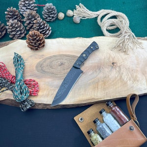 May include: A black and silver knife with a textured blade rests on a wooden surface. The knife is accompanied by a leather pouch containing small glass bottles. Also present are pine cones, rope, and colorful cords.