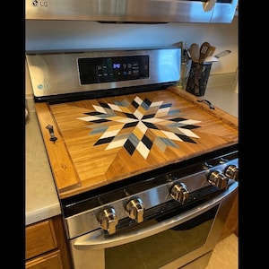 May include: A wooden stovetop cover with a geometric starburst design in black, white, blue, and beige. The cover is placed on a stainless steel stovetop with four knobs.