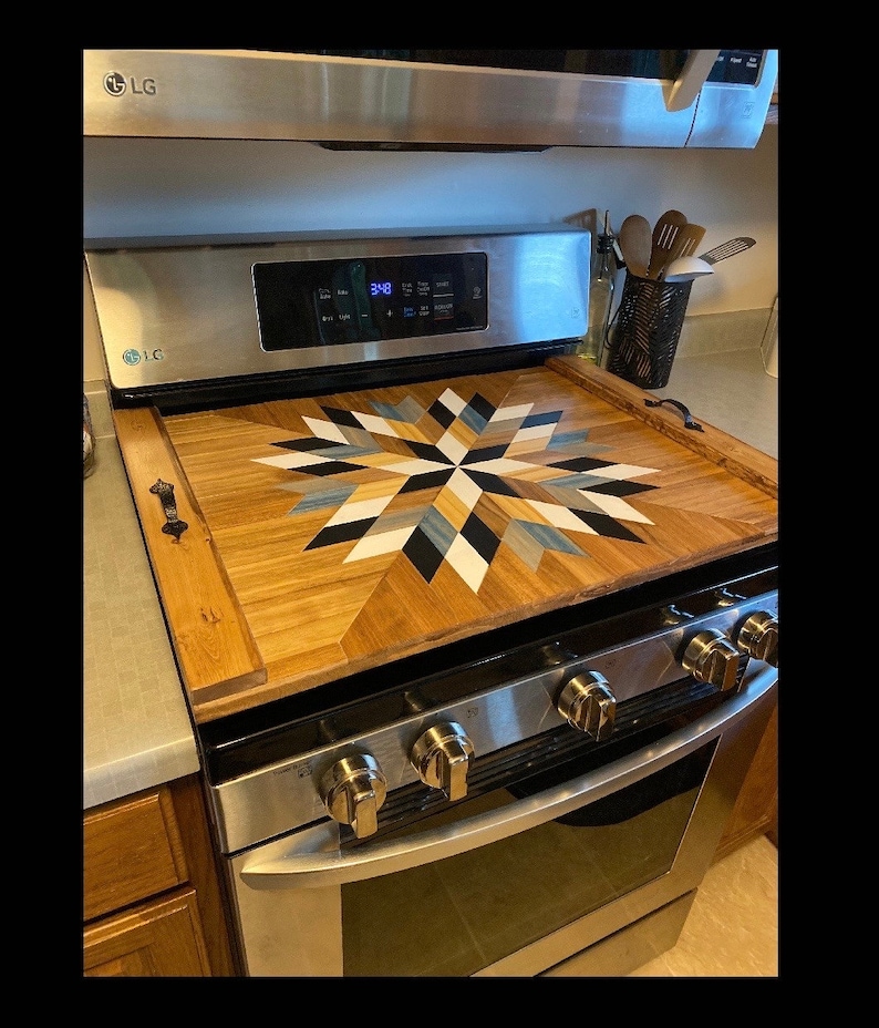 May include: A wooden stovetop cover with a geometric starburst design in black, white, blue, and beige. The cover is placed on a stainless steel stovetop with four knobs.
