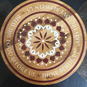May include: A round wooden serving tray with a brown and white mandala design. The tray has a white inscription that reads "And the love between us. Food before us. Those beside us. Bless this".