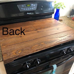 May include: A wooden stovetop cover with a dark brown stain. The cover is sitting on top of a black stove with a digital clock display that reads 2:33. The word "Back" is written on the cover.