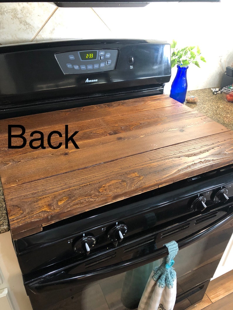 May include: A wooden stovetop cover with a dark brown stain. The cover is sitting on top of a black stove with a digital clock display that reads 2:33. The word "Back" is written on the cover.