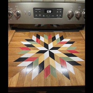 May include: A wooden stovetop cover with a starburst design in black, white, red, green, and tan. The cover is made of wood and has a glossy finish.