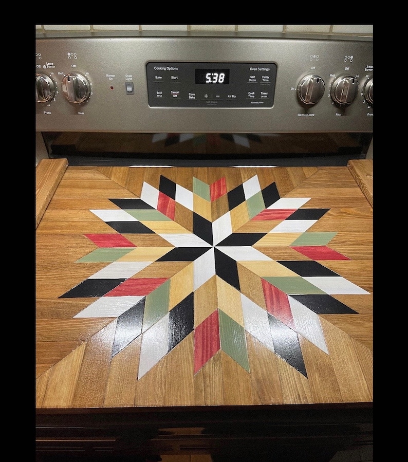 May include: A wooden stovetop cover with a starburst design in black, white, red, green, and tan. The cover is made of wood and has a glossy finish.