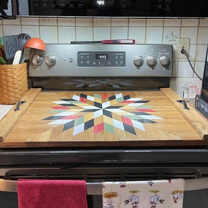 May include: A wooden stovetop cover with a colorful quilt pattern. The cover is made of wood and has a black handle on each side. The cover is sitting on top of a black stovetop.