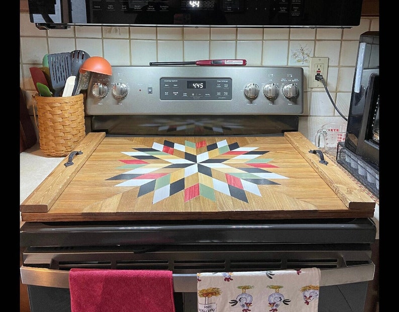 May include: A wooden stovetop cover with a colorful quilt pattern. The cover is made of wood and has a black handle on each side. The cover is sitting on top of a black stovetop.