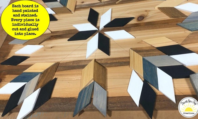 May include: Close-up of a wooden wall art piece with a geometric design. The piece is made of various wood pieces, painted in black, white, and shades of blue and brown. The text "Each board is hand painted and stained. Every piece is individually cut and glued into place." is visible in the image.
