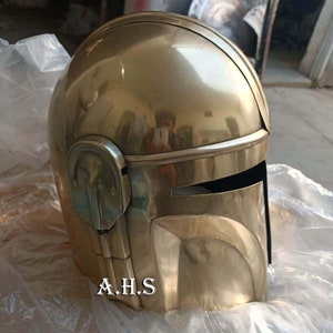 May include: A gold-colored helmet with a visor and a rounded top. The helmet has a metallic sheen and is resting on a white surface. The text "A.H.S" is printed on the lower front of the helmet.