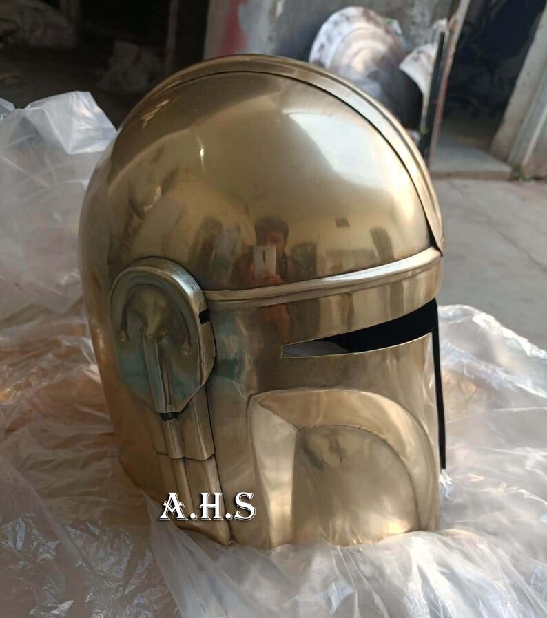 May include: A gold-colored helmet with a visor and a rounded top. The helmet has a metallic sheen and is resting on a white surface. The text "A.H.S" is printed on the lower front of the helmet.