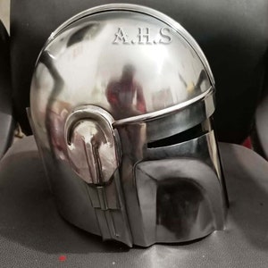 May include: A polished, metallic helmet with a visor and distinctive angular features. The helmet has a smooth, reflective surface, with the letters "A.H.S" etched on the top. The design suggests a futuristic or science fiction theme.