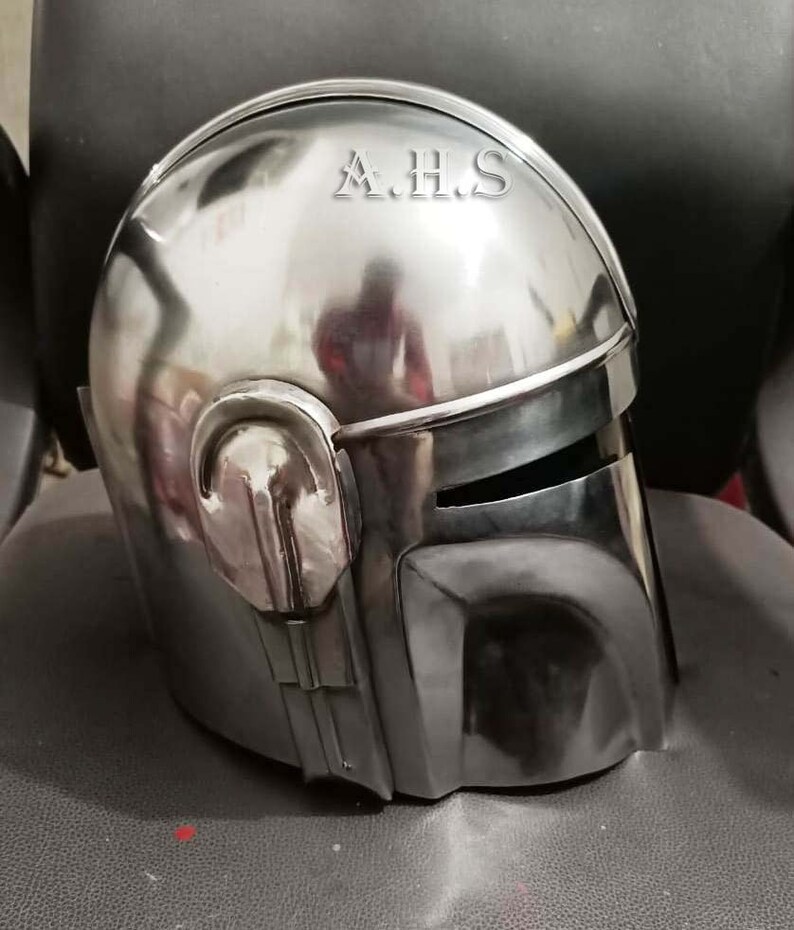 May include: A polished, metallic helmet with a visor and distinctive angular features. The helmet has a smooth, reflective surface, with the letters "A.H.S" etched on the top. The design suggests a futuristic or science fiction theme.