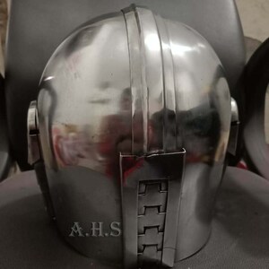 May include: A polished, silver-colored helmet with vertical ridges on top and a rectangular plate on the back. The initials "A.H.S" are visible on the lower back of the helmet. The helmet is likely for cosplay or historical reenactment.