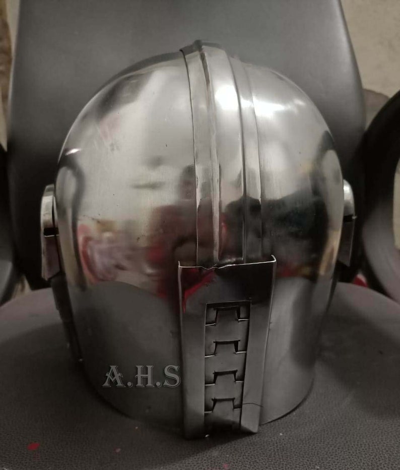 May include: A polished, silver-colored helmet with vertical ridges on top and a rectangular plate on the back. The initials "A.H.S" are visible on the lower back of the helmet. The helmet is likely for cosplay or historical reenactment.