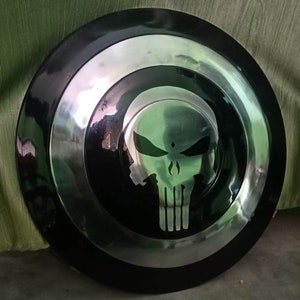 May include: A black and silver round shield with a white skull graphic in the center. The skull has a white outline and is set against a black background.