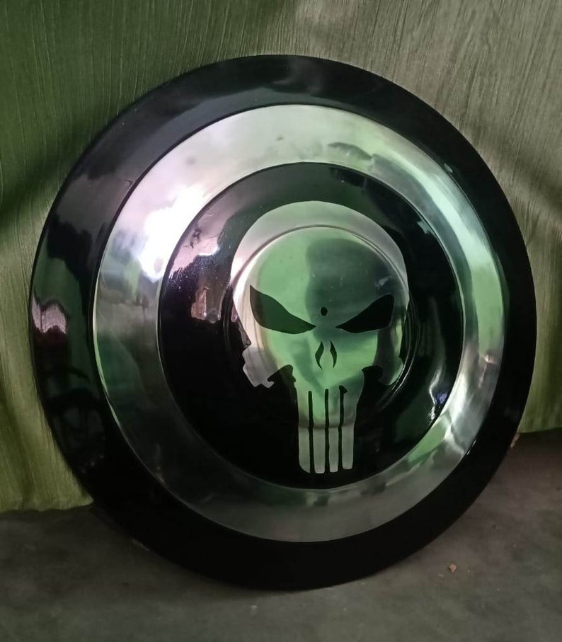 May include: A black and silver round shield with a white skull graphic in the center. The skull has a white outline and is set against a black background.