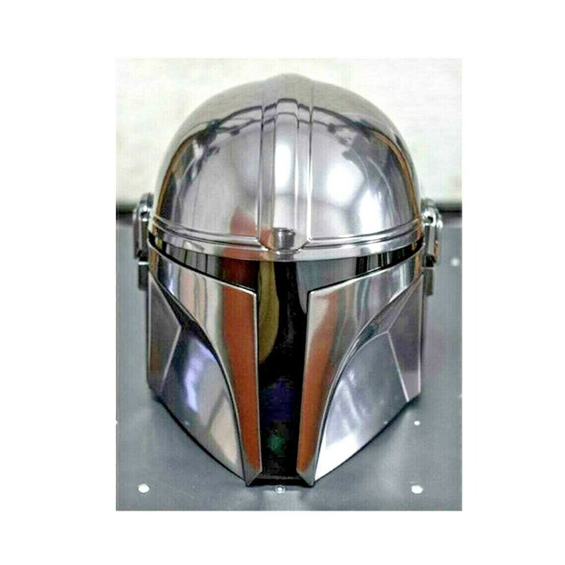 May include: A shiny, silver Mandalorian helmet. The helmet has a rounded top with vertical ridges and a distinctive visor. The metallic surface reflects light, creating highlights and shadows. The helmet is a replica from the Star Wars franchise.