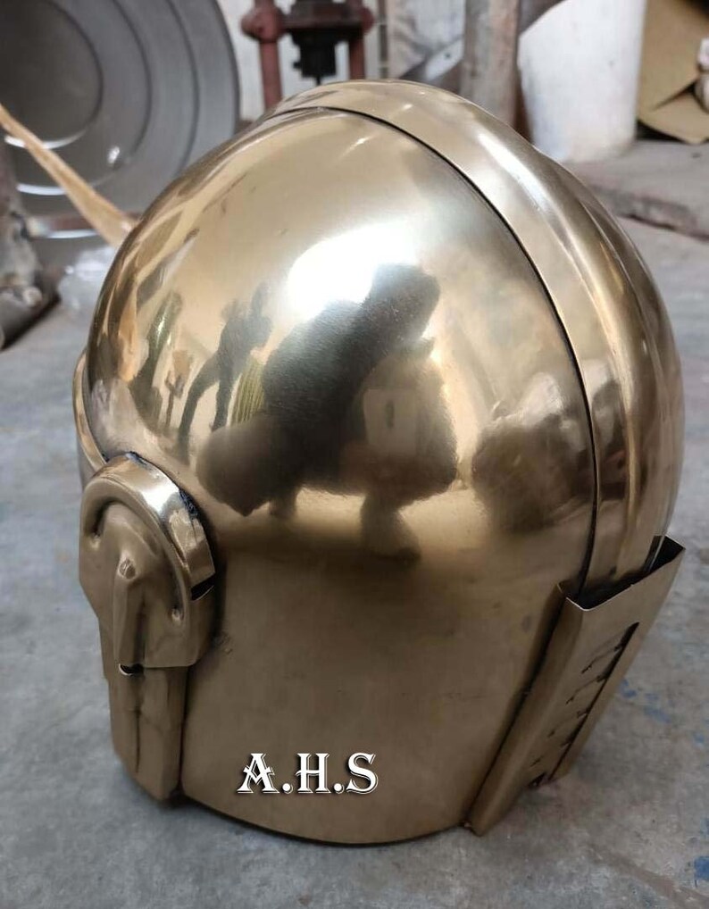May include: A polished brass helmet with a rounded top and a detailed face guard. The helmet has a reflective surface, and the initials "A.H.S" are visible on the lower front. The design suggests a historical or fantasy theme.
