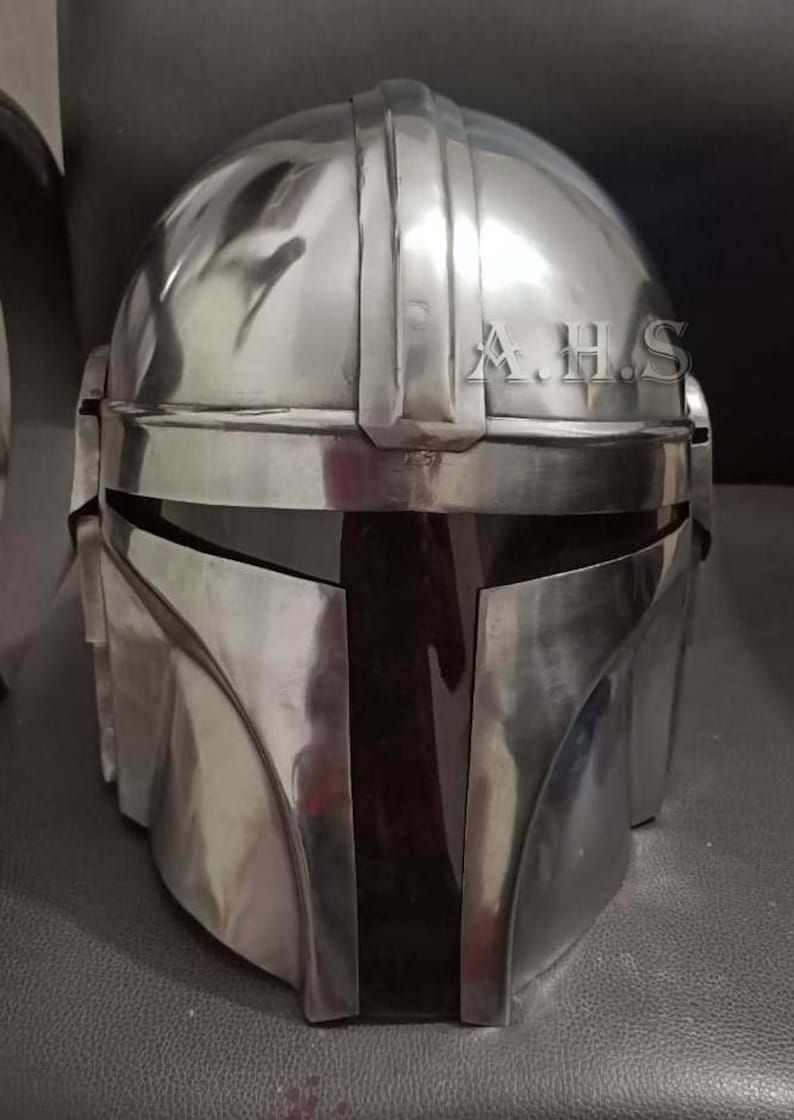 May include: A silver-colored helmet with a visor and a rounded top. The helmet has vertical ridges on the top and the letters "A.H.S" are visible on the side. The helmet is likely a prop or costume piece.