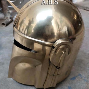 May include: A gold-colored helmet with the text "A.H.S" on the top. The helmet has a smooth, reflective surface and features a visor and ear pieces. The design is reminiscent of a space-age or futuristic aesthetic.