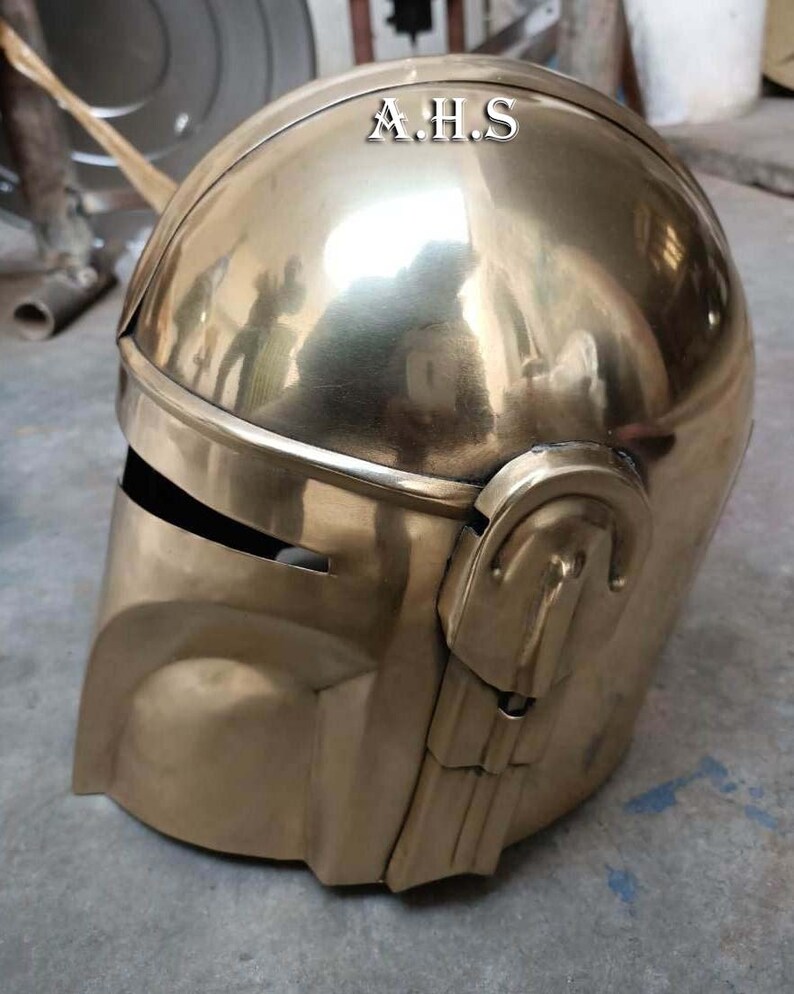 May include: A gold-colored helmet with the text "A.H.S" on the top. The helmet has a smooth, reflective surface and features a visor and ear pieces. The design is reminiscent of a space-age or futuristic aesthetic.