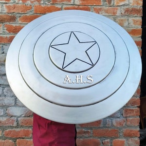 Peut inclure: Un bouclier de Captain America argent&eacute; avec une &eacute;toile noire au centre. Le bouclier a trois cercles concentriques autour de l'&eacute;toile. Le bouclier est tenu devant un mur de briques. Le texte "A.H.S." est imprim&eacute; sur le bouclier.
