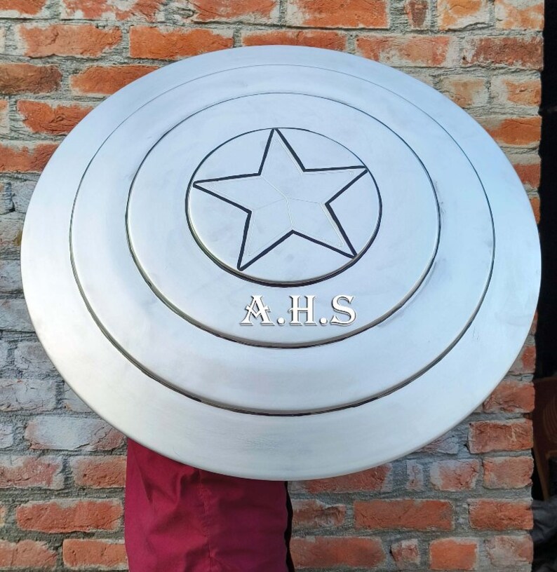 Peut inclure: Un bouclier de Captain America argent&eacute; avec une &eacute;toile noire au centre. Le bouclier a trois cercles concentriques autour de l'&eacute;toile. Le bouclier est tenu devant un mur de briques. Le texte "A.H.S." est imprim&eacute; sur le bouclier.