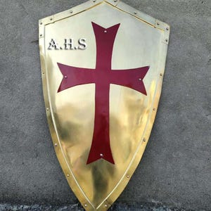 May include: A polished brass heater shield featuring a large, deep red cross centered on the surface. The shield is bordered by a metal rim secured with small rivets. The letters A.H.S are clearly visible in silver on the upper left side.