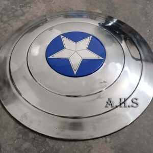 Pu&ograve; includere: Uno scudo di Captain America in metallo argentato con una stella blu e bianca al centro. Lo scudo ha un bordo rialzato ed &egrave; leggermente ammaccato. Il testo "A.H.S." &egrave; visibile in basso a destra dello scudo.