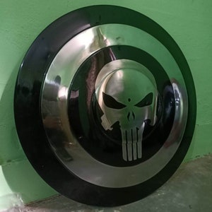May include: A black and silver round shield with a white skull graphic in the center. The skull has a black outline and is stylized to look like the Punisher logo.