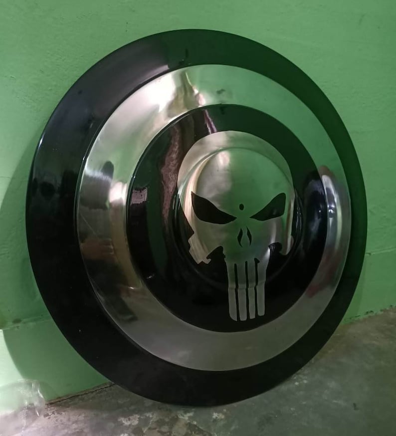 May include: A black and silver round shield with a white skull graphic in the center. The skull has a black outline and is stylized to look like the Punisher logo.