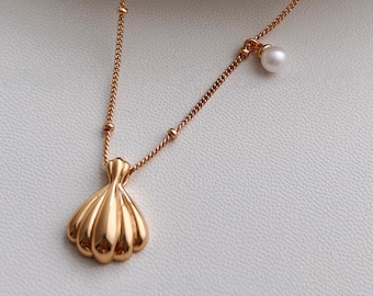 Op de afbeelding: Een goudkleurige ketting met een schelpenhanger en een kleine parelbedel. De ketting heeft kleine, met kralen bezette accenten. De schelpenhanger is gedetailleerd met gebogen ribbels. De parel is wit.