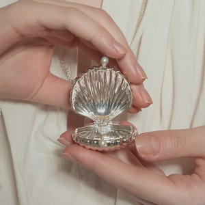 Op de afbeelding: Een zilveren schelpenhangerketting, open om een parel te onthullen. De hanger wordt opengehouden en toont de ingewikkelde details van het schelpenontwerp. De ketting staat op een witte achtergrond.