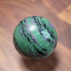 May include: A polished spherical stone featuring a vibrant green base color with prominent streaks of deep black and patches of magenta-pink mineral inclusions throughout the surface, creating a natural marbled appearance on the round object.