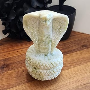 May include: A carved stone cobra statue with a flared hood and detailed scales. The statue is light green and white, set on a textured base. A black decorative tree and a black abstract sculpture are in the background.