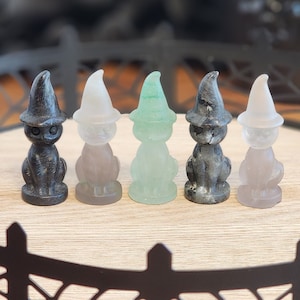 May include: Five small black cat figurines wearing witch hats. The hats are in different colors: black, white, green, and gray. The figurines are sitting on a black tray with a spiderweb design.
