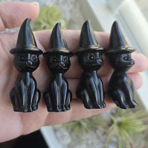 May include: Four black obsidian witch cat figurines wearing black pointed hats. The figurines are carved in a sitting position with their paws tucked under them.