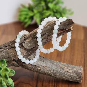 May include: White beaded bracelets displayed on a piece of driftwood. The bracelets are made of round, opaque beads. The background includes a wooden surface and green plants, creating a natural aesthetic. The bracelets are a simple, elegant accessory.