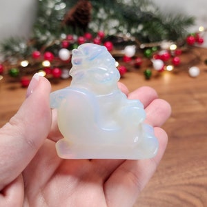 May include: A white opalite Christmas sleigh figurine with a white opalite Santa Claus figure sitting inside. The figurine is about 2 inches tall and is held in a person's hand.