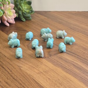 May include: A collection of twelve small, hand-carved animal figurines made from light blue, semi-translucent stone. Each figurine features a rounded body, short legs, and a snout, depicting a pig-like creature arranged on a wooden surface.
