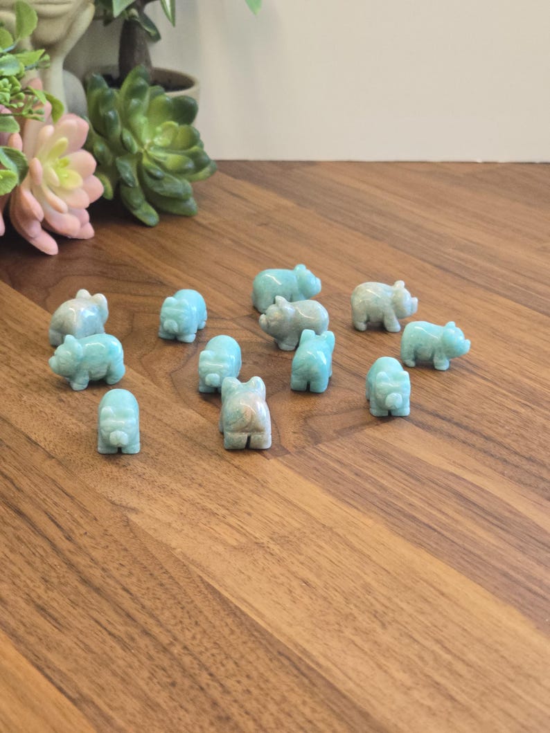 May include: A collection of twelve small, hand-carved animal figurines made from light blue, semi-translucent stone. Each figurine features a rounded body, short legs, and a snout, depicting a pig-like creature arranged on a wooden surface.