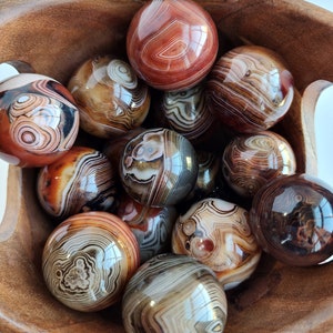 May include: A wooden bowl filled with agate spheres. The spheres are various shades of brown, red, and white with intricate banding patterns.