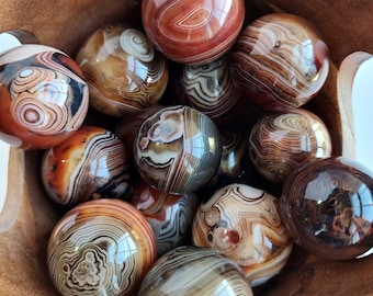 Op de afbeelding: Een houten kom gevuld met agaat bollen. De bollen zijn in verschillende tinten bruin, rood en wit met ingewikkelde bandpatronen.
