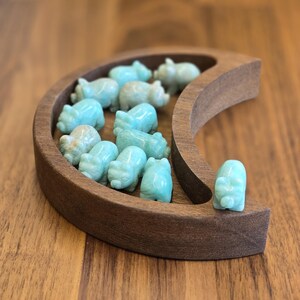 May include: A crescent moon-shaped wooden tray containing eleven small, carved light blue stone pig figurines. The tray has a smooth, dark brown finish, and the stone pigs are arranged within the curved interior and on the outer edge.