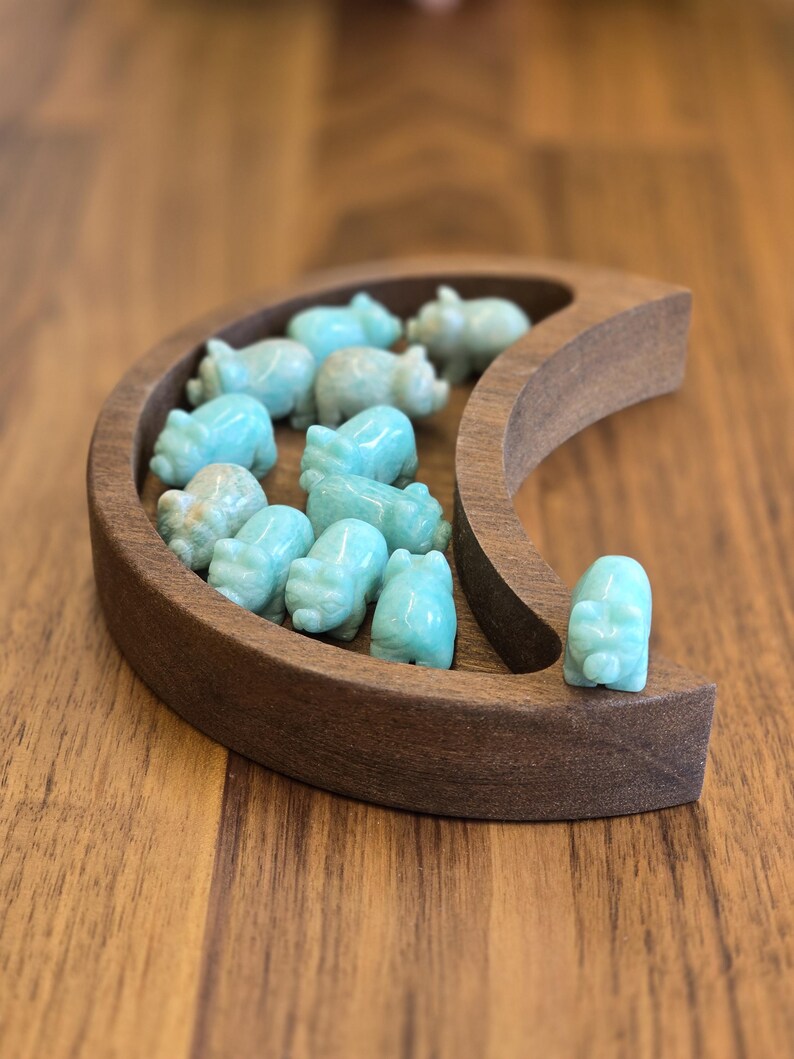 May include: A crescent moon-shaped wooden tray containing eleven small, carved light blue stone pig figurines. The tray has a smooth, dark brown finish, and the stone pigs are arranged within the curved interior and on the outer edge.