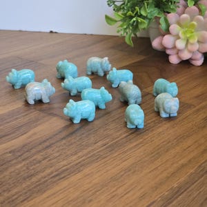 May include: A collection of twelve small, hand-carved animal figurines made from light blue, opaque stone. Each figurine is shaped like a pig with distinct ears, a snout, and four legs, arranged in a scattered formation across the surface.